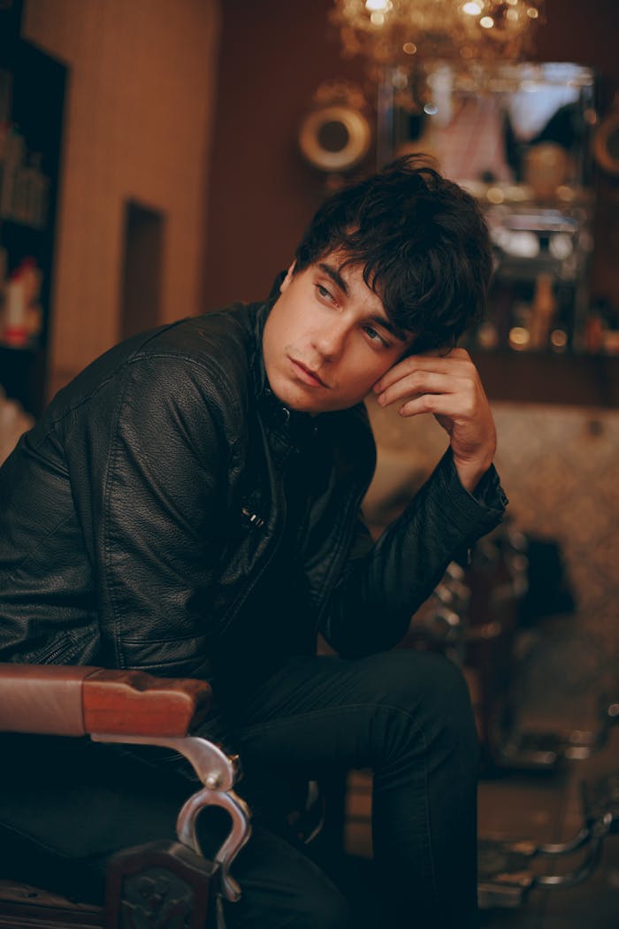 A young man seated indoors wearing a black leather jacket, showcasing a contemplative mood.