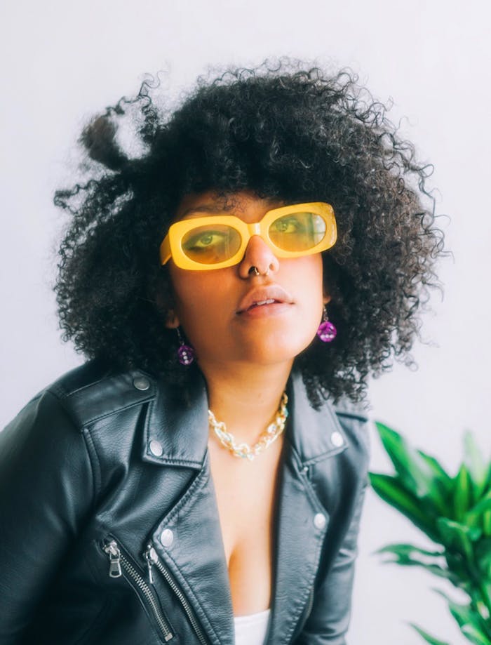 Stylish young woman in sunglasses and leather jacket posing with vibrant hair and accessories.