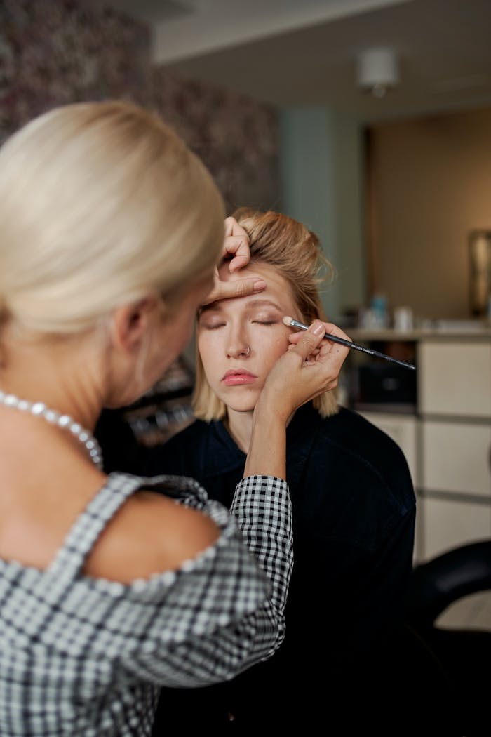 A makeup artist applies eyeshadow to a young woman in a serene, indoor salon setting.