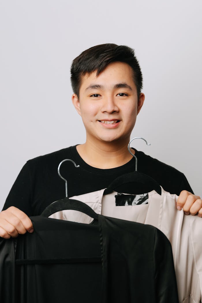 about-us A young East Asian man smiling while holding clothing hangers against a plain white background.